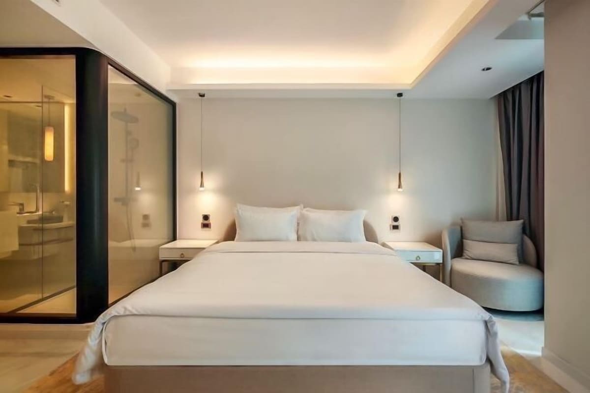 Une chambre moderne avec un grand lit, des tables de chevet, un fauteuil et une cloison en verre.