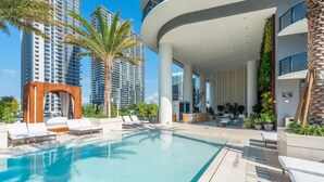 Ein moderner Hotelpoolbereich mit Liegestühlen, einer Bogenstruktur aus Stein und hoch aufragenden Gebäuden im Hintergrund.