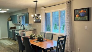 A dining area with a wooden table and chairs, a kitchen in the background, and a window with curtains.