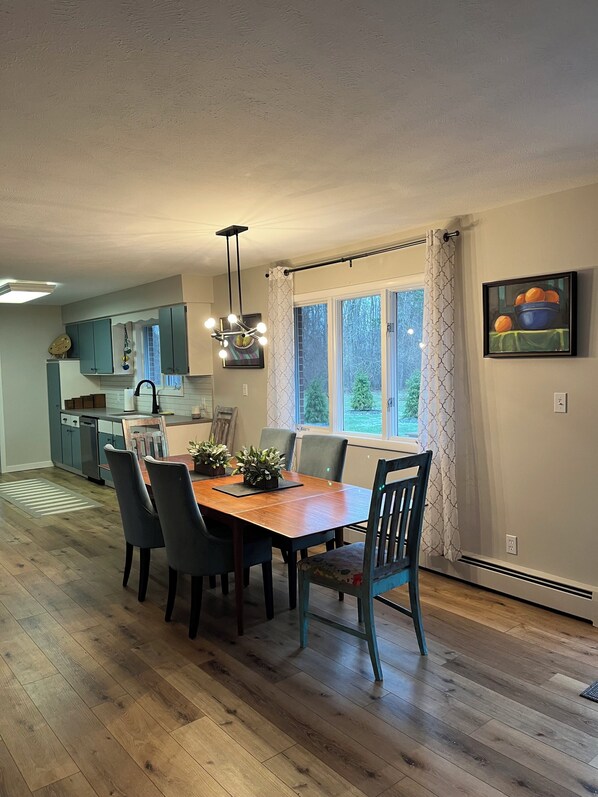 A dining area with a wooden table and chairs, a kitchen in the background, and a window with curtains.
