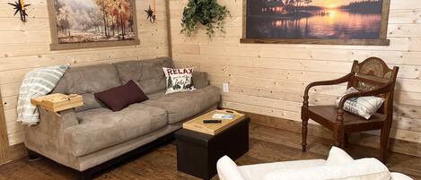 Un interior de cabaña acogedor con un sofá gris, un sillón blanco, una mesa de centro de madera y dos cuadros que representan paisajes tranquilos.