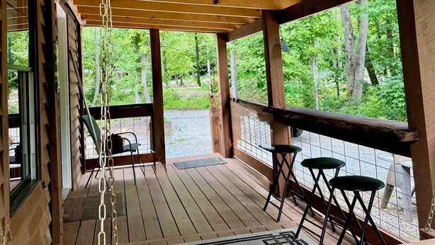 A wooden deck with a swing chair, a table, and a view of a forest. - Charming 2-bedroom cabin in beautiful mountains of Pickens, SC. (Pickens)