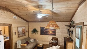 A cozy cabin interior with a wooden ceiling, a ceiling fan, a sofa, a chair, a small table, and a window with a view of a sunset. - Charming 2-bedroom cabin in beautiful mountains of Pickens, SC. (Pickens)