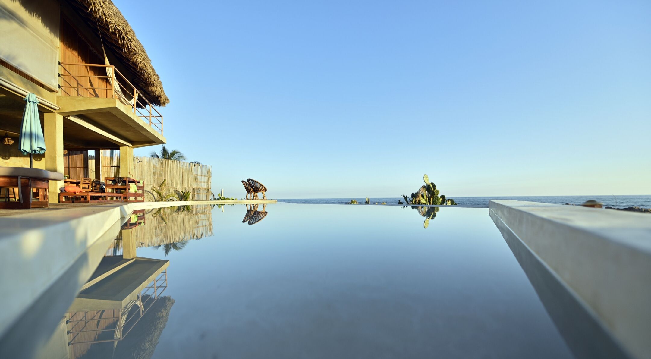 Una piscina infinita con vistas claras al océano, una estructura de techo de paja y muebles de madera.