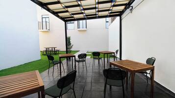 A covered outdoor patio with tables and chairs, a small lawn, and a building in the background.