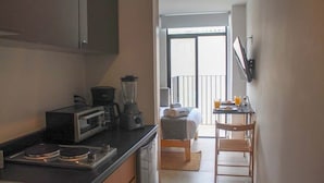 Cozinha compacta com torradeira, liquidificador e cafeteira. Pequena área de jantar com mesa e cadeira visíveis, que dá para uma varanda com vista.