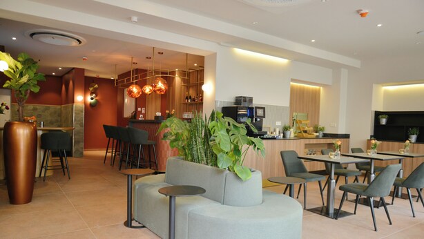 A modern hotel lobby with seating areas, a bar, and a kitchen in the background. - Day8 Hotel  (Cologne)