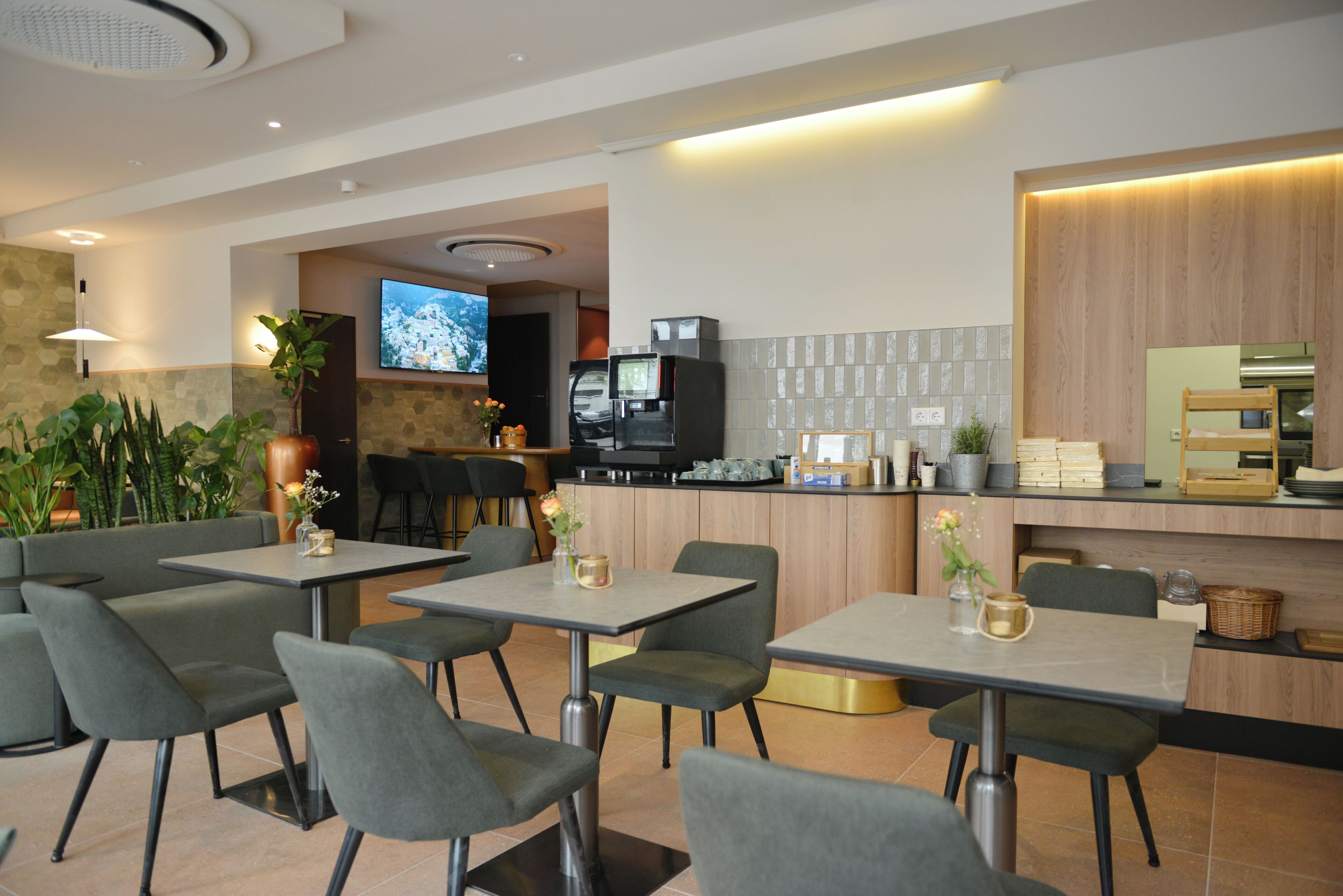 A modern restaurant with grey upholstered chairs and tables, a TV screen, and a kitchen area with wooden cabinets.
