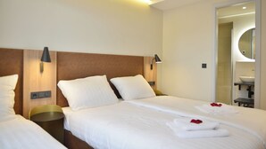 A hotel room with two beds, a wooden headboard, bedside lamps, a bathroom with a sink and mirror, and a shower. - Day8 Hotel  (Cologne)