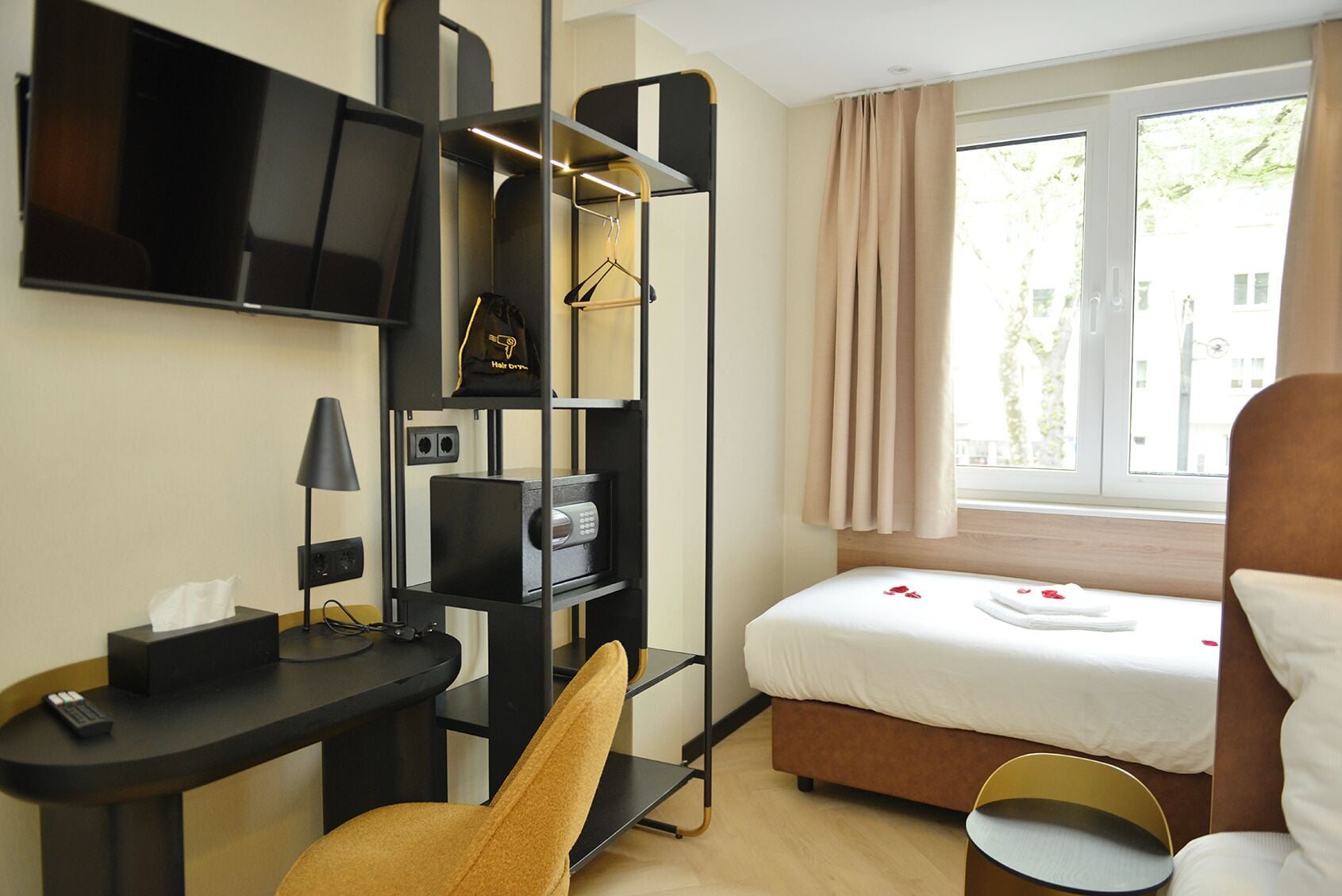 Una camera d'albergo con un letto, una scrivania, una sedia, una TV, uno scaffale con un asciugacapelli e una finestra con tende.