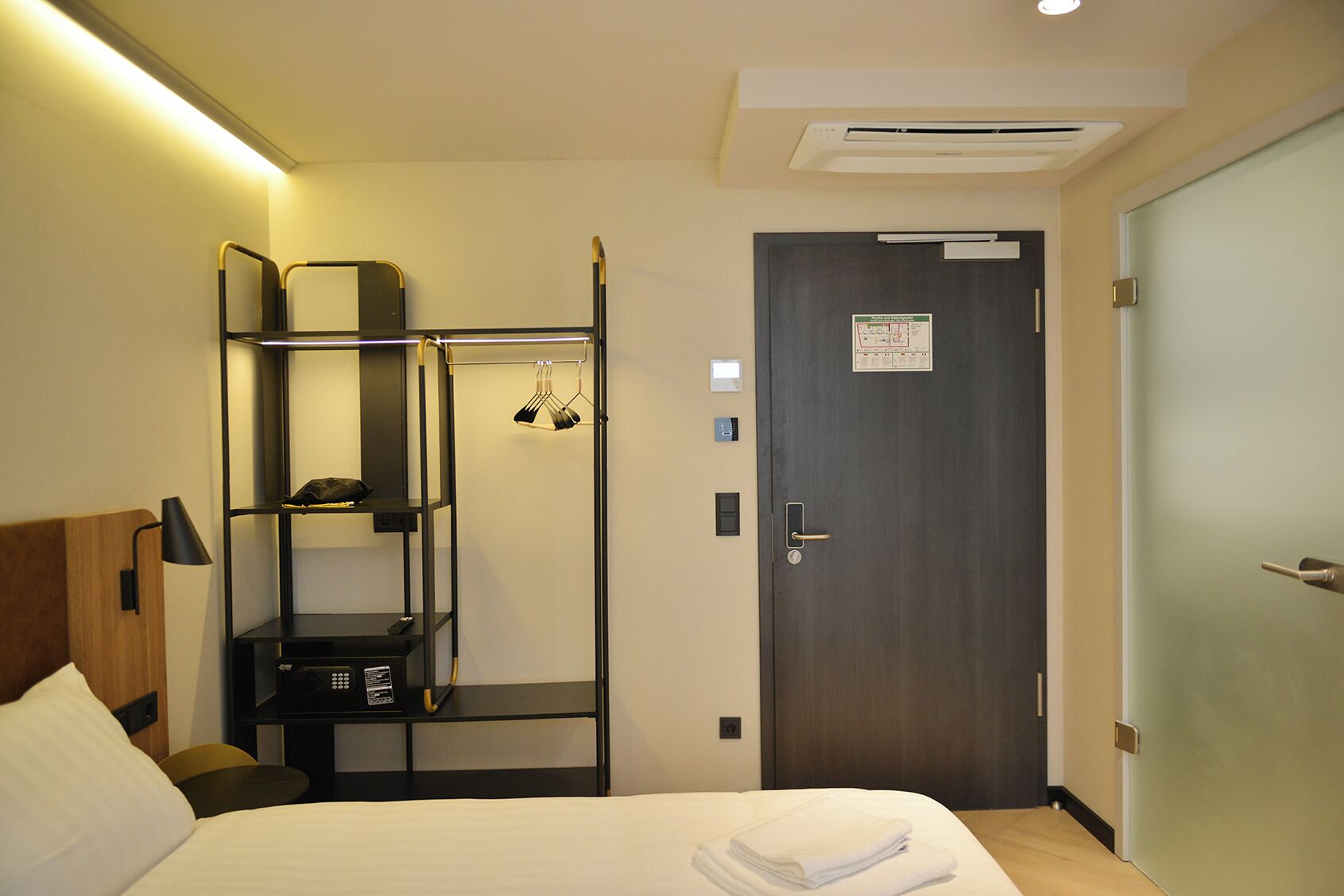 A hotel room with a bed, a bedside table, a wardrobe, a shelf unit, and a door with a sign.