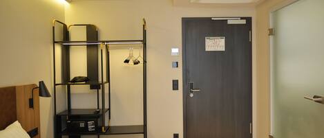 A hotel room with a bed, a bedside table, a wardrobe, a shelf unit, and a door with a sign.