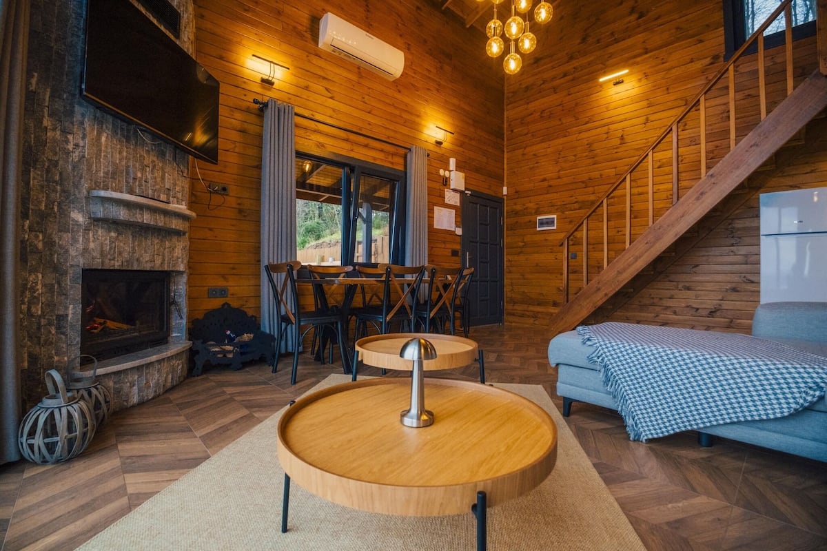 A cozy cabin interior with a fireplace, wooden floors, a dining area, and a staircase.