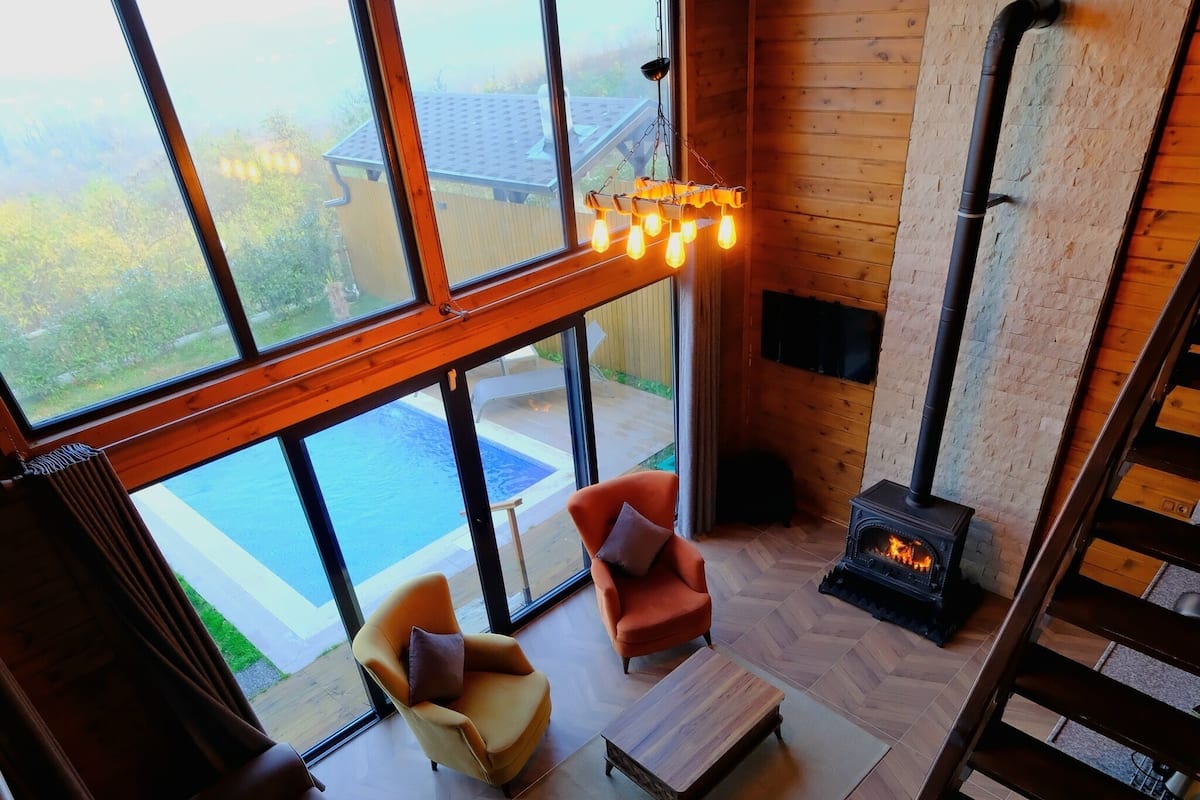 A cozy room with a large window, a sofa, a chair, a fireplace, and a wooden staircase.
