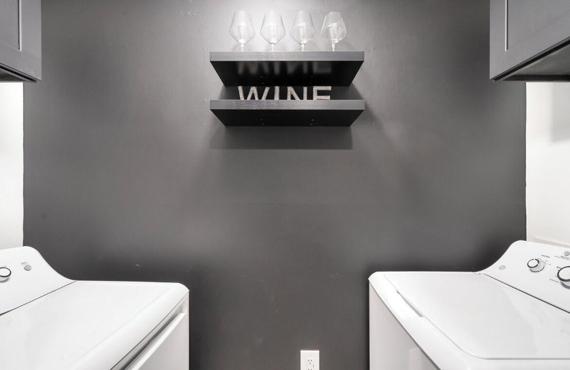 A modern laundry area with two washing machines, a wine rack with glasses, and a dark wall.