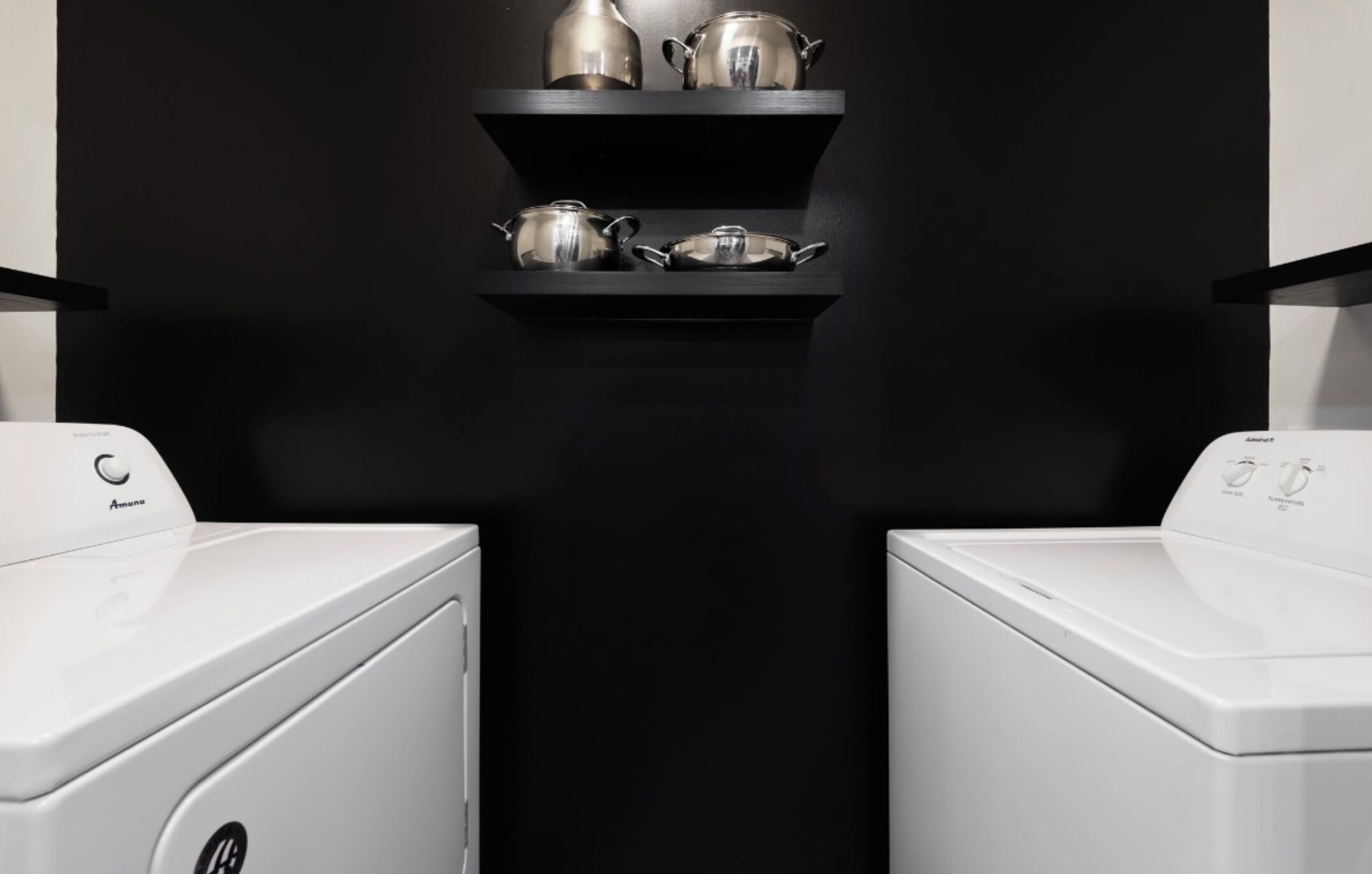 A laundry area with two white washing machines and a black shelf above them holding silver kitchenware.