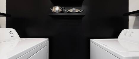 A laundry area with two white washing machines and a black shelf above them holding silver kitchenware.