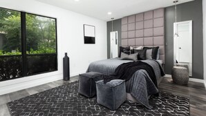 Ein modernes Schlafzimmer mit großem Fenster, einem Bett mit grauer Bettwäsche, einem grauen Kopfteil und einem schwarz-weißen Teppich.