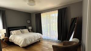 A modern bedroom with a large bed, a wooden floor, a brown armchair, and a window with curtains. - Serene Farm Stay (Nanaimo)