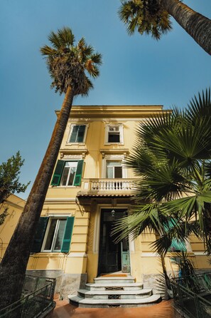 A two-story building with a balcony, a staircase leading to the entrance, and a palm tree in the foreground. - CasaChi | View of Capri, a balcony, and charm in the heart of Chiaia—Naples (Napoli)