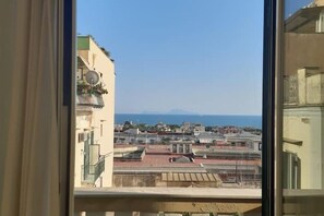 A view from a window overlooking a coastal cityscape with buildings and a distant mountain. - CasaChi | View of Capri, a balcony, and charm in the heart of Chiaia—Naples (Napoli)