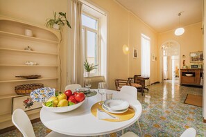 A dining area with a table set for two, a bowl of fruit, and a window with curtains.