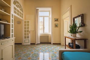 A room with a tiled floor, a radiator, a television on a cabinet, a plant on a side table, and a bed with a blue cover.
