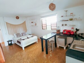 Ein kleiner Raum mit Bett, Tisch, Hocker, Küchenzeile und Fenster.