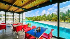A poolside dining area with red chairs and a table, overlooking a pool and palm trees. - Cana Brava Residences | Panoramic Condo | Golf View (Punta Cana)