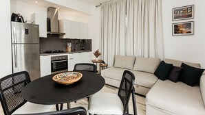 A modern kitchen with a refrigerator, stove, and cabinets. There is a dining area with a table and chairs. A sofa is against the wall, and curtains are drawn. - Cana Brava Residences | Panoramic Condo | Golf View (Punta Cana)