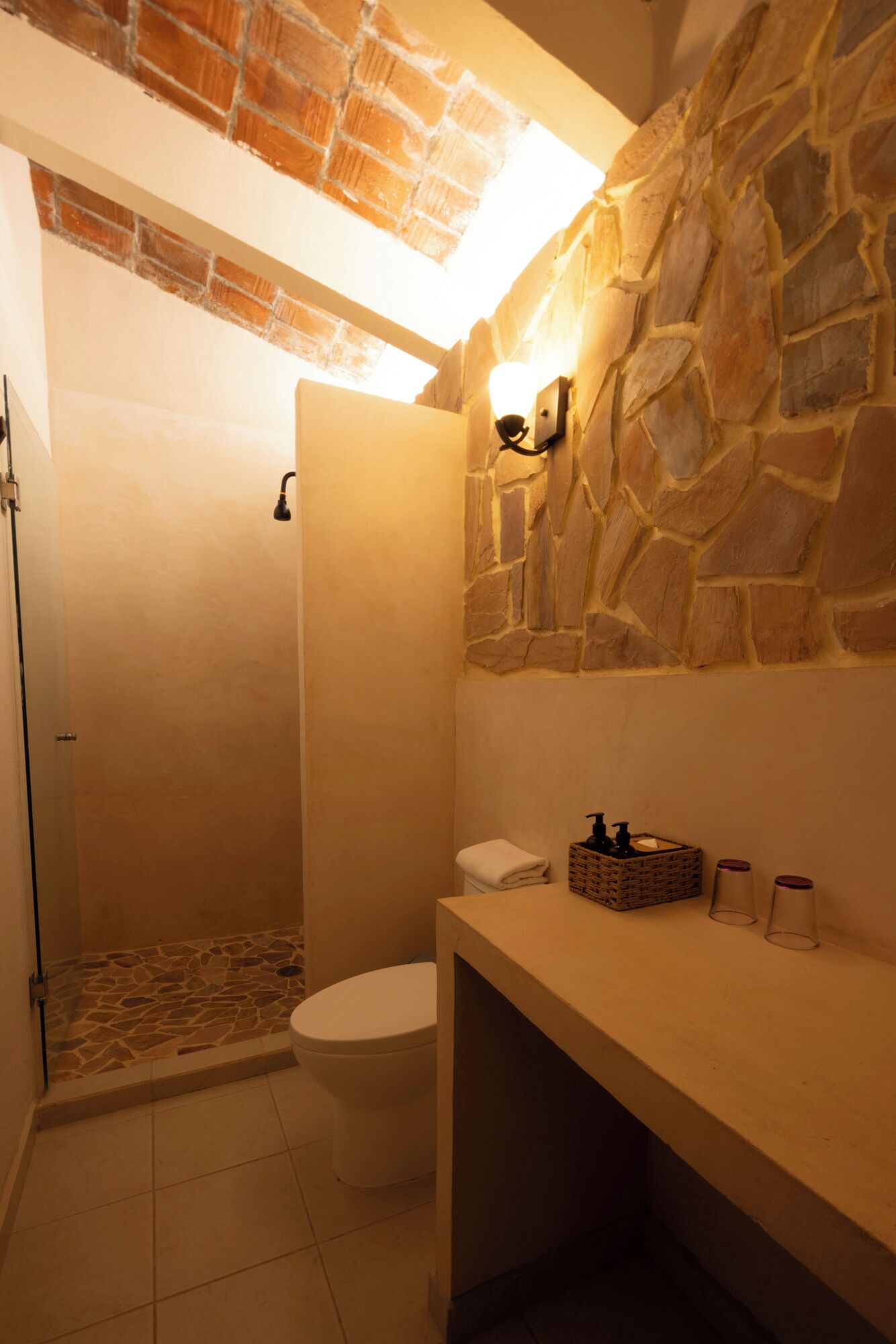 A bathroom with a stone wall, a shower, a toilet, and a sink area with toiletries.