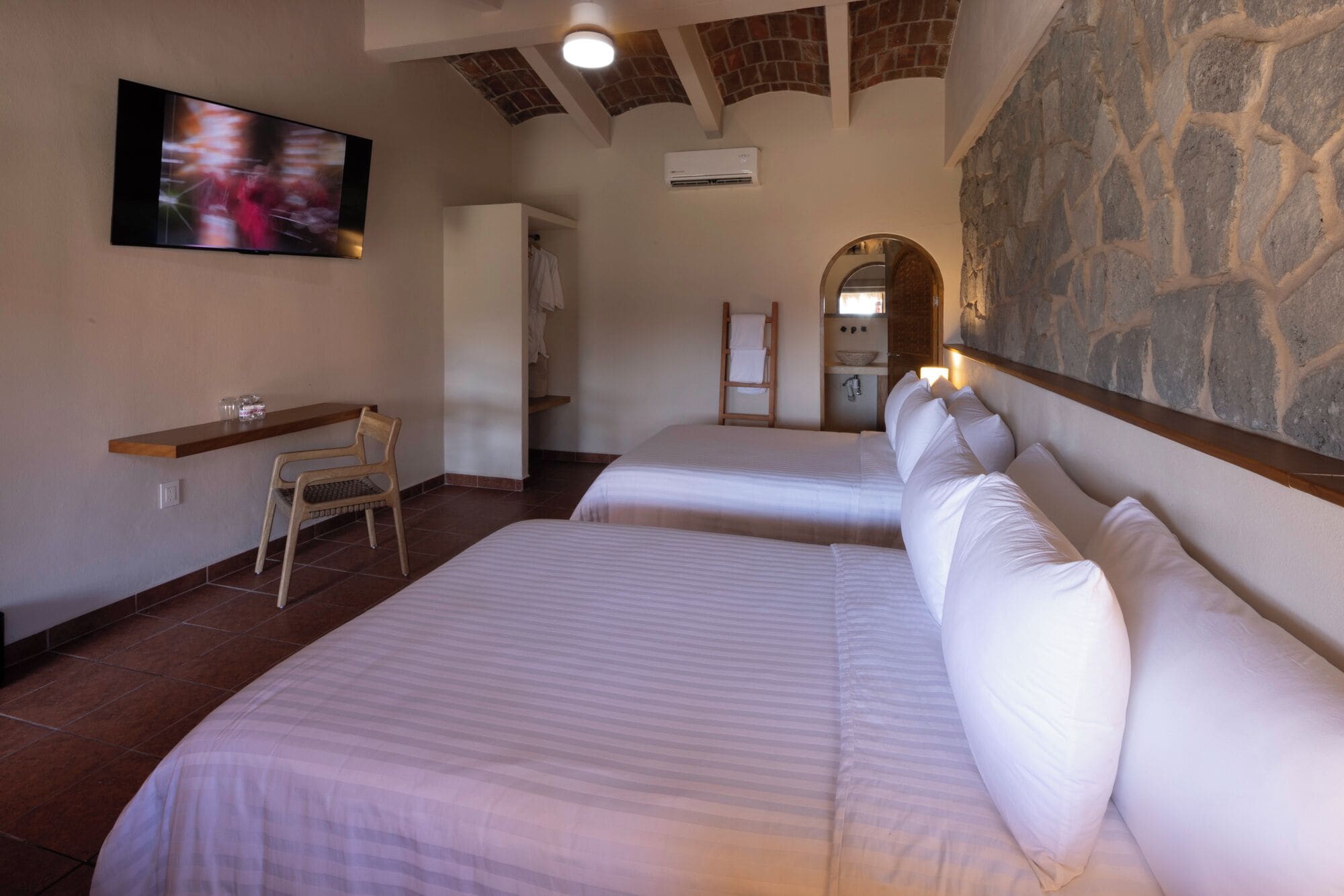 A hotel room with two beds, a TV, a wooden chair, and a stone wall.