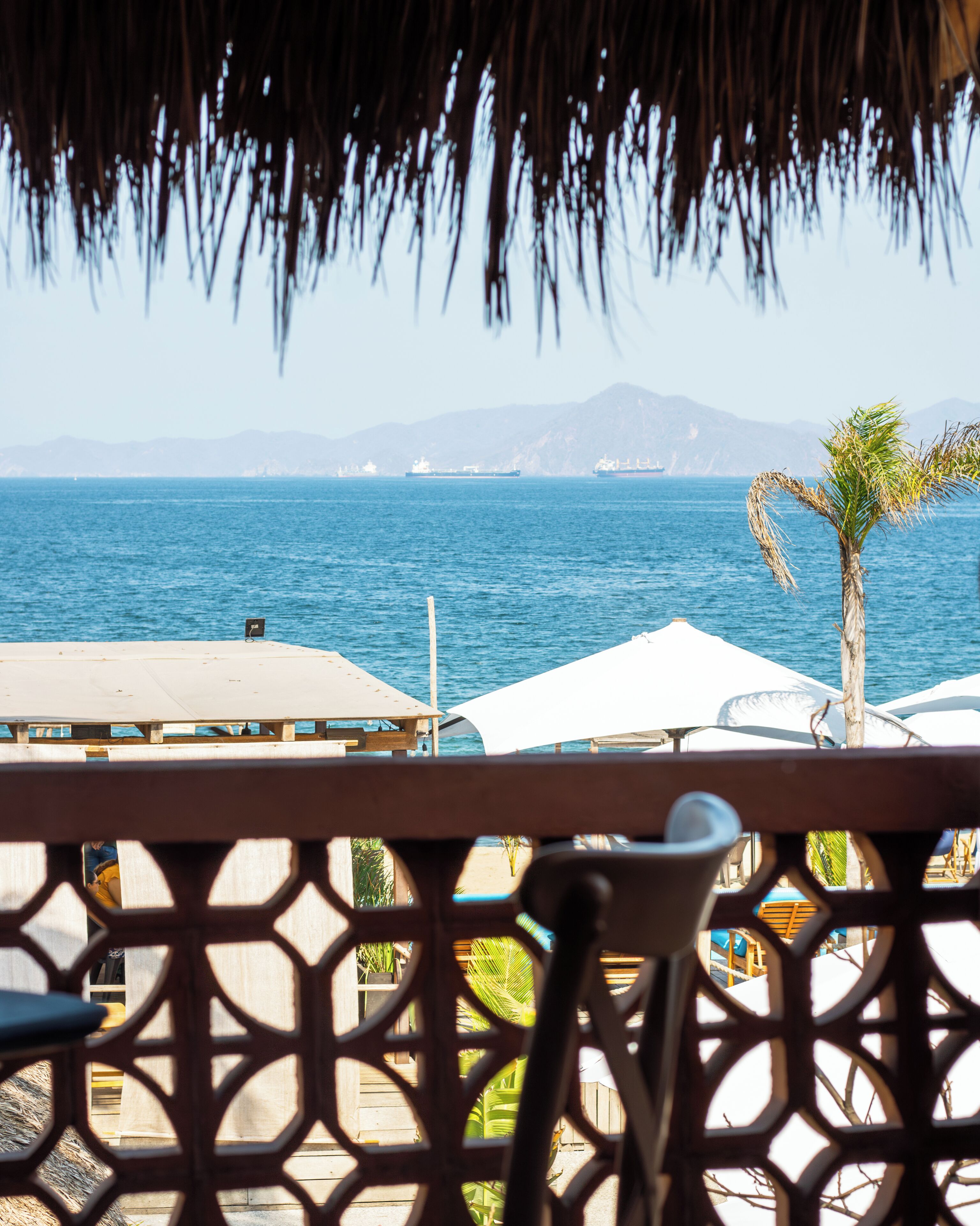 A view from a covered outdoor area with a thatched roof, overlooking a calm sea and distant mountains.