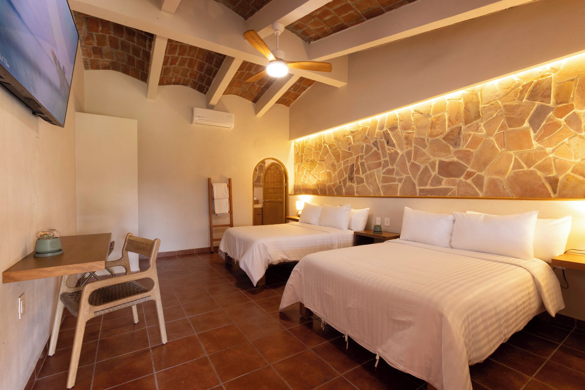 Habitación de hotel con dos camas, un escritorio, una silla, un televisor y un muro de piedra.