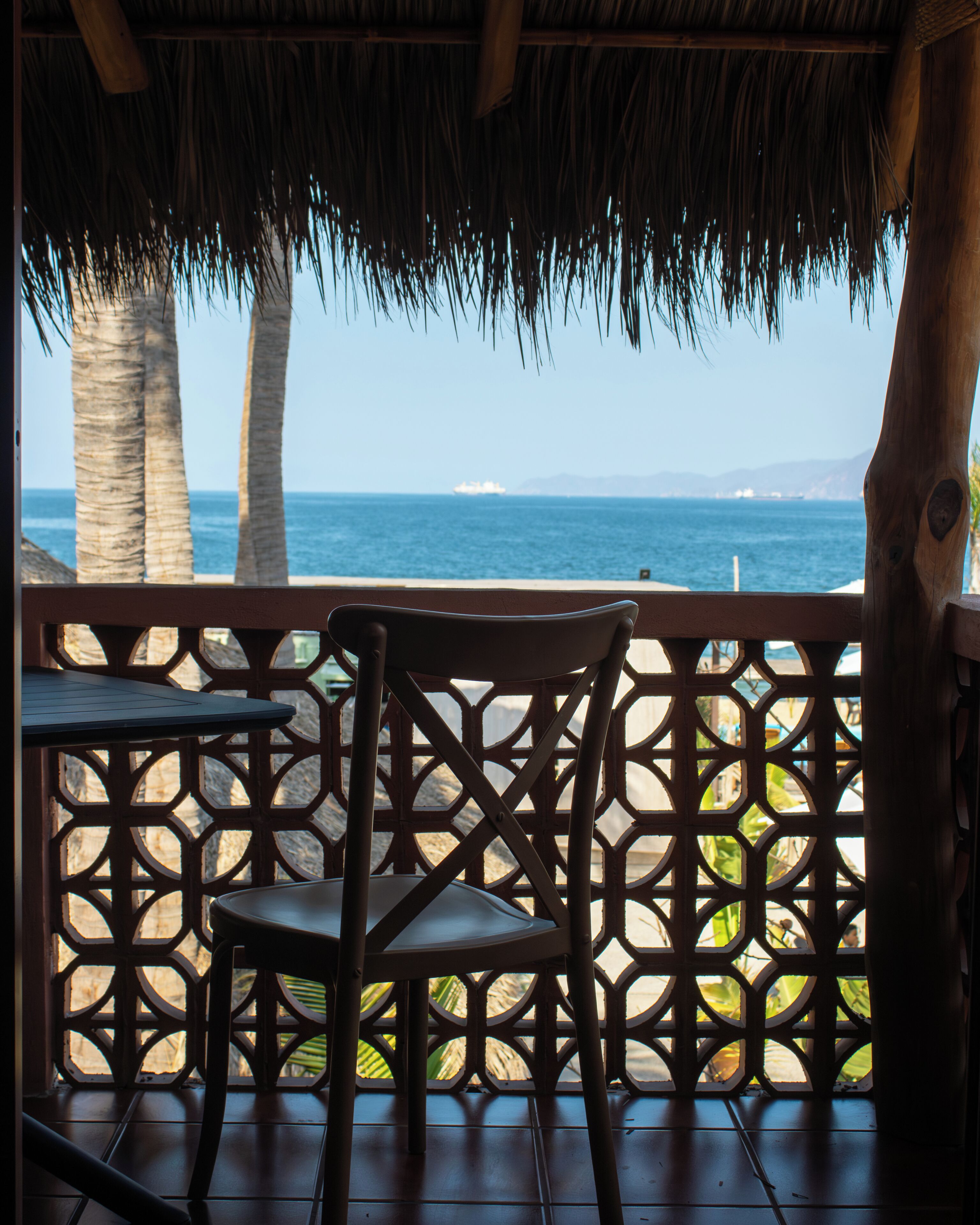 A wooden chair and table on a balcony with a view of the ocean and palm trees.