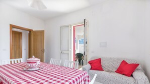 A room with a checkered table, white chairs, a sofa with red cushions, and a door leading to another room.