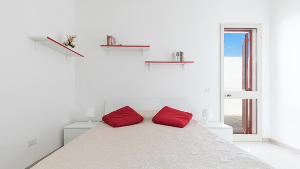 A bedroom with a bed, two red pillows, a nightstand, a white lamp, and three white shelves with red edges against a white wall.