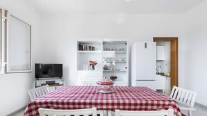 A dining area with a checkered tablecloth, white chairs, a TV, and a kitchen area with shelves and a refrigerator.