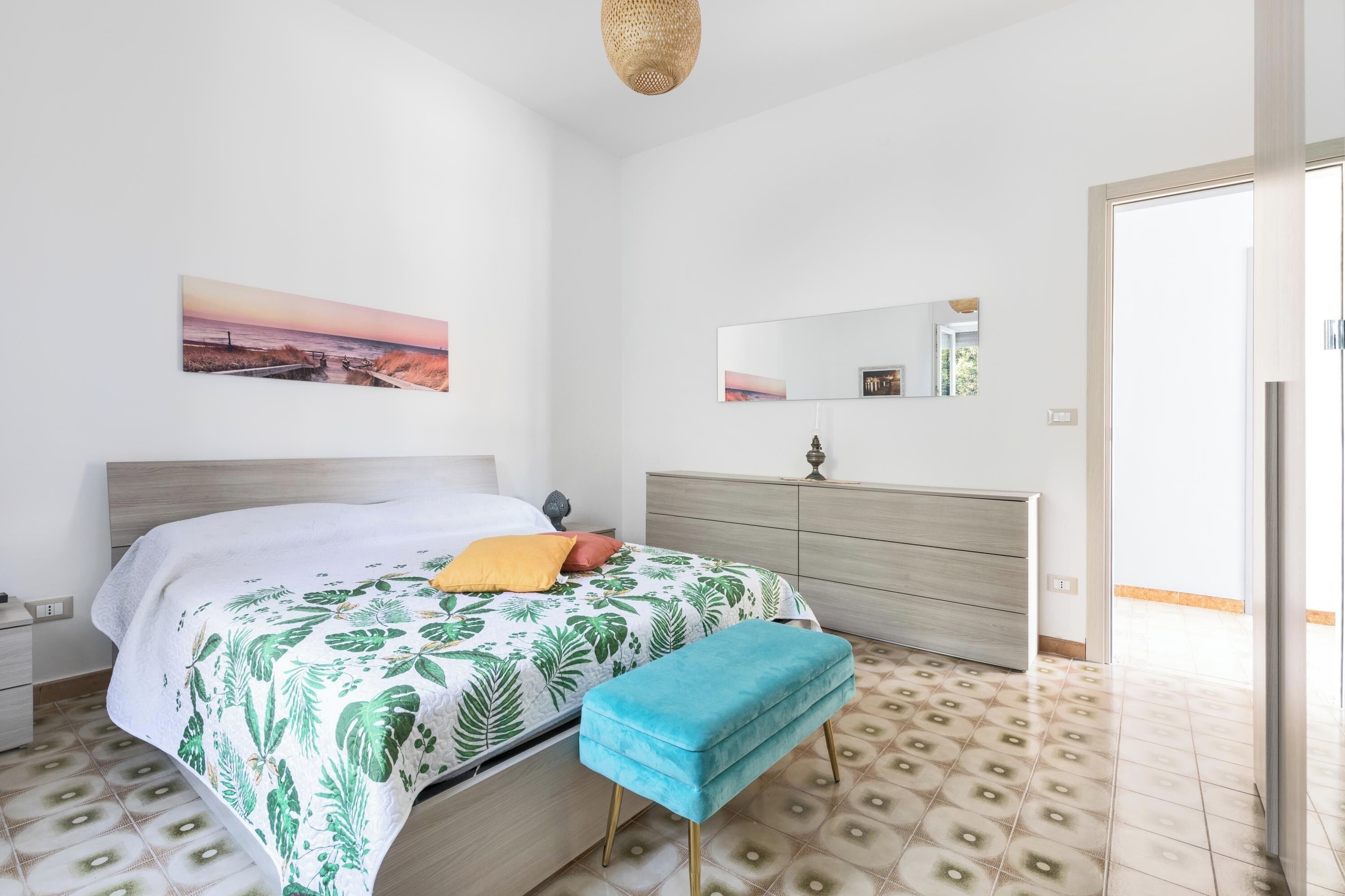 Ein Schlafzimmer mit Bett, einer Bank, einem Schminktisch, einem Spiegel und einer Tür zu einem anderen Raum.