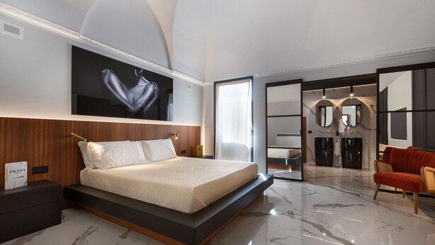 A modern bedroom with a large bed, a flat-screen TV, a wardrobe, and a seating area with a red sofa.
