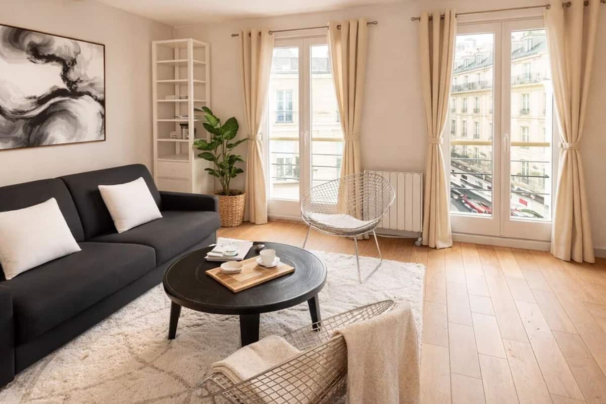 A modern living room with a black sofa, a round coffee table, a white chair, and a large window with curtains.