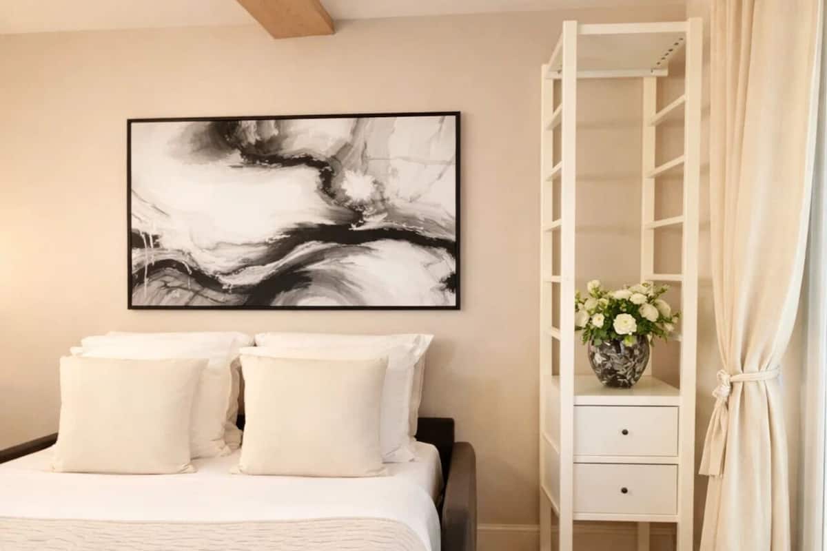 A bedroom with a bed, a bedside table with a vase of flowers, a wardrobe, and a large abstract painting on the wall.