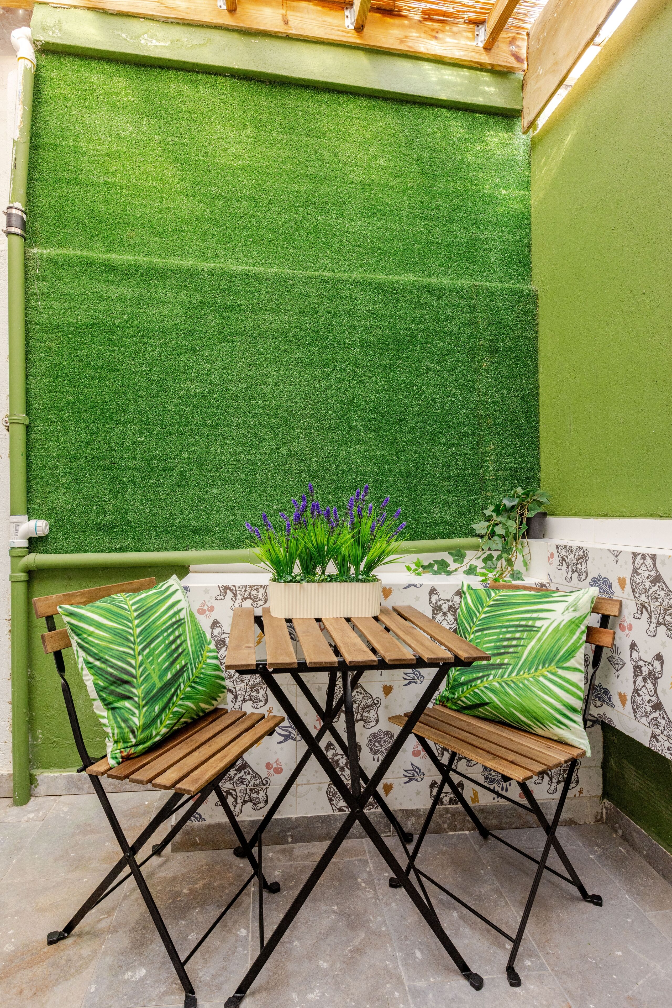 Un petit espace extérieur aménagé avec une table en bois et deux chaises, entouré de gazon synthétique vert et doté d’un sol carrelé.