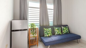 A modern living room with a grey sofa, a small wooden table with a plant, and a white refrigerator. - City Beach Apartments (San Juan)