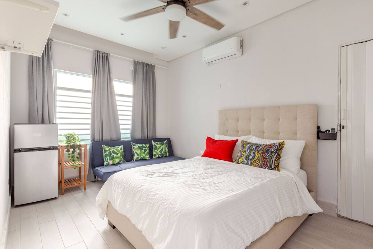 Un dormitorio con cama, ventilador de techo, una kitchenette pequeña y una ventana con cortinas.