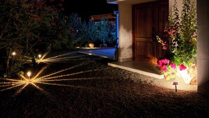 A modern building with a well-lit entrance, surrounded by landscaped gardens with flowering plants and a pathway illuminated by star-shaped lights.