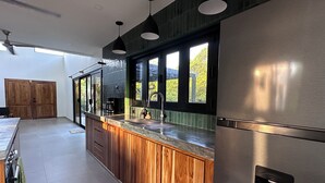 A modern kitchen with wooden cabinets, a stainless steel refrigerator, and a large window providing a view of greenery outside.