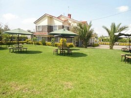 Un espacio al aire libre con césped, mesas, sombrillas, una casa al fondo y palmeras.
