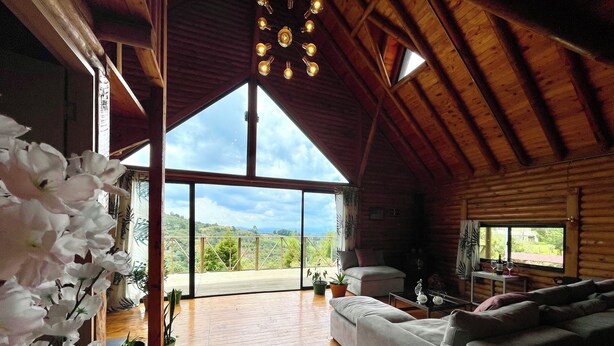 A spacious room with a large wooden ceiling, a grey sofa, a dining table with wine glasses, and a balcony with a view of the landscape.
