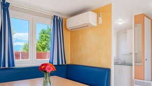 Una habitación pequeña con una mesa de madera, un banco acolchado azul, un florero con flores y un equipo de aire acondicionado.